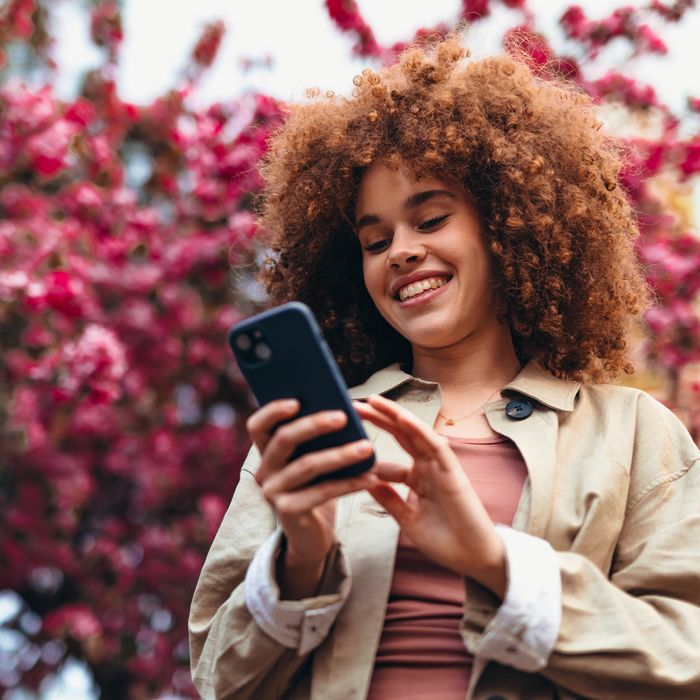 Smiling young woman with curly hair enjoying online communication while using a smartphone, surrounded by a blooming pink tree in spring, embracing the beauty of nature in the parkColorful Human