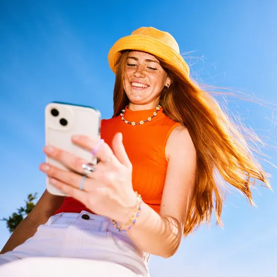 Woman in her 20s reading messages on cell phone with ahppy expression, against clear blue sky, low angle viewColorful Human