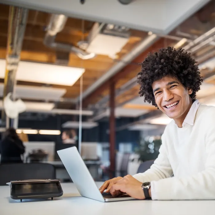 Portrait of happy young man sitting at table with laptop looking at camera and smiling. Egyptian businessman in casuals working on laptop computer.