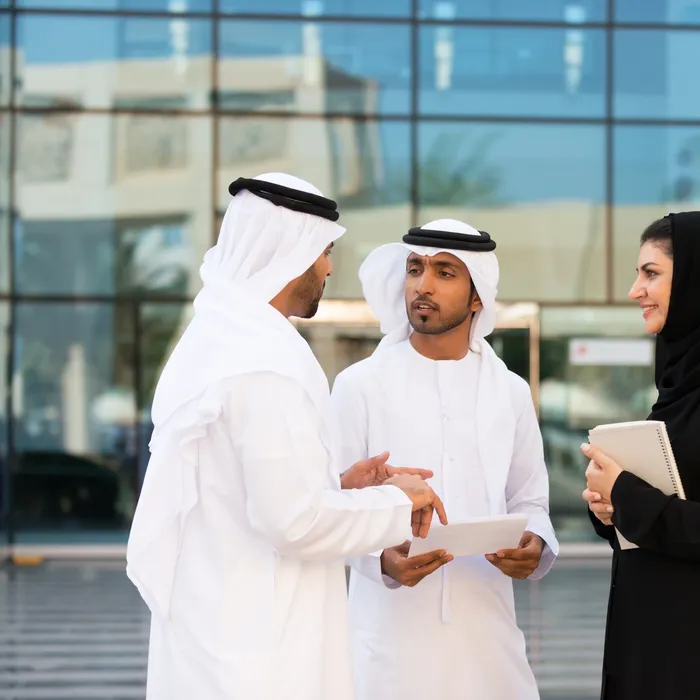 Dubai iStockalypse.  Traditionally dressed young adult Arab and Emirati men and woman talking outside a modern office building.  Businesswoman is wearing traditional abaya and hijab. Businessmen are wearing kandoora, agal and ghutra.  Holding notebooks, documents, contracts.  Modern blue glass office building and palm trees reflected in the background.  Dubai, UAE, Middle East.