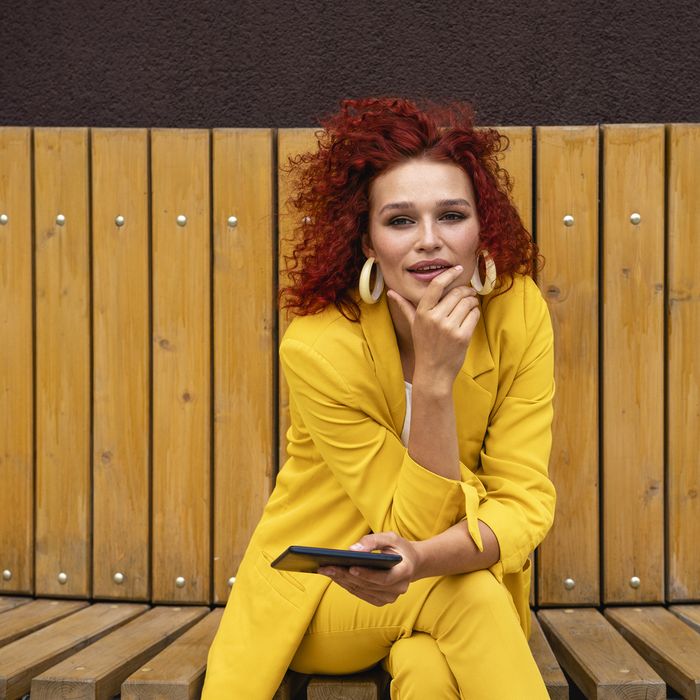 Young woman with curly hair and yellow suit, sitting on bench, using digital tablet