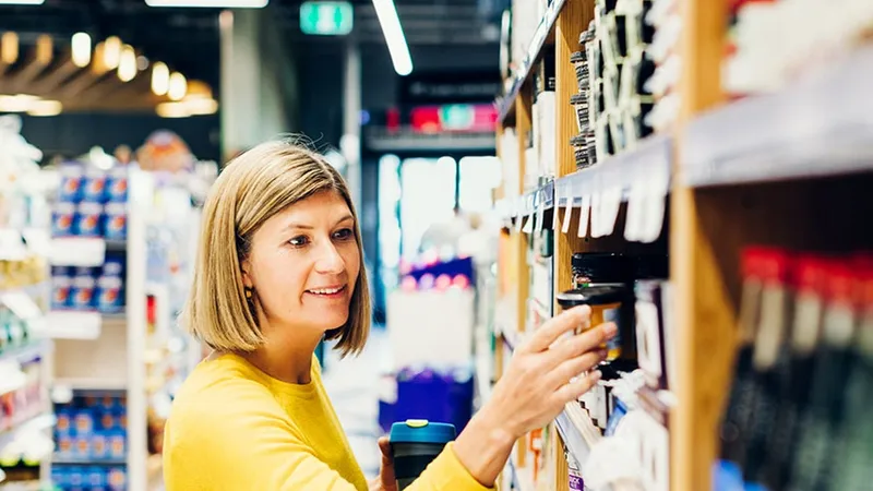 A woman in a yellow blouse picks up a product from a shelf in a grocery store