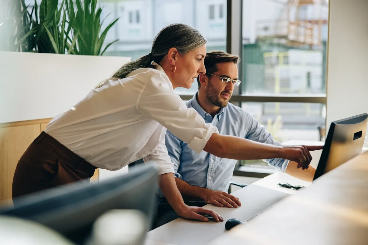 Mature woman working with young man sitting at desk and showing something on computer screen in office. Female executive pointing at desktop monitor and talking with male colleague in office.
