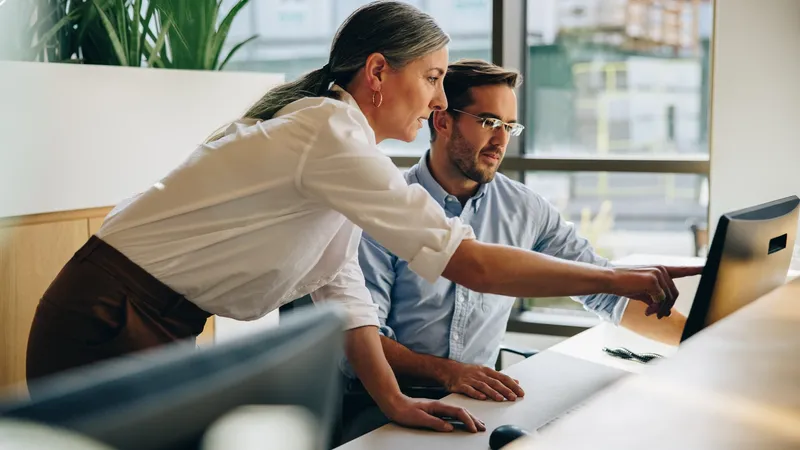 Mature woman working with young man sitting at desk and showing something on computer screen in office. Female executive pointing at desktop monitor and talking with male colleague in office.