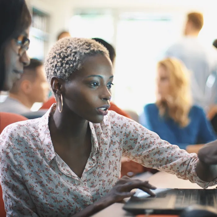 Closeup side view of two mid 20's  black women doing their design project on a computer. They might be software developers as well.  Blurry coworkers in background, all released.
