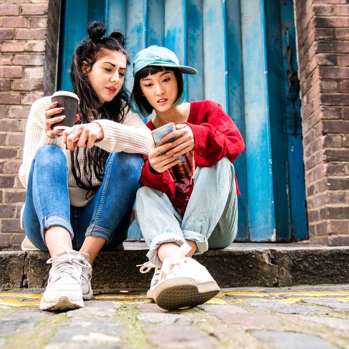 Two young women sitting on sidewalk looking at smartphone