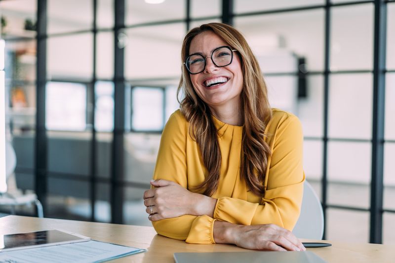Portrait of a beautiful smiling businesswoman sitting on desk in her office and looking at camera.
