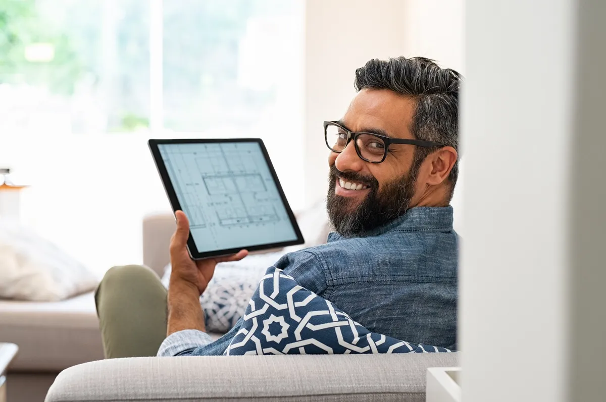 Senior architect working on blueprint for client on digital tablet while relaxing at home. Architect drawing blueprint on digital tablet using software while lying on couch. Rear view of confident customer checking house project layout on laptop at home while looking at camera.