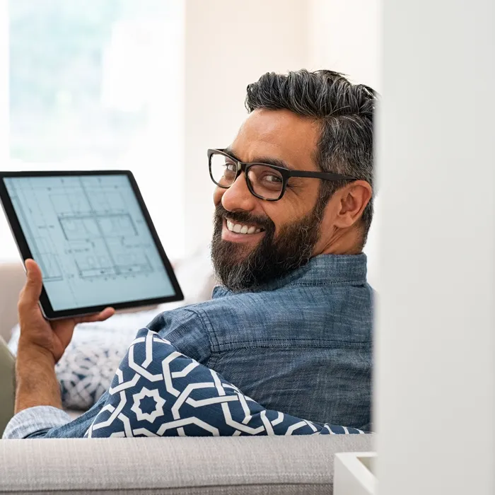 Senior architect working on blueprint for client on digital tablet while relaxing at home. Architect drawing blueprint on digital tablet using software while lying on couch. Rear view of confident customer checking house project layout on laptop at home while looking at camera.