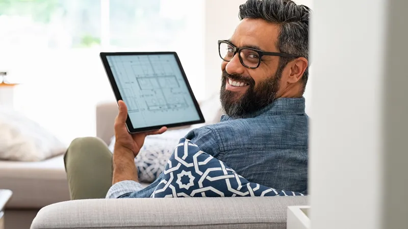 Senior architect working on blueprint for client on digital tablet while relaxing at home. Architect drawing blueprint on digital tablet using software while lying on couch. Rear view of confident customer checking house project layout on laptop at home while looking at camera.