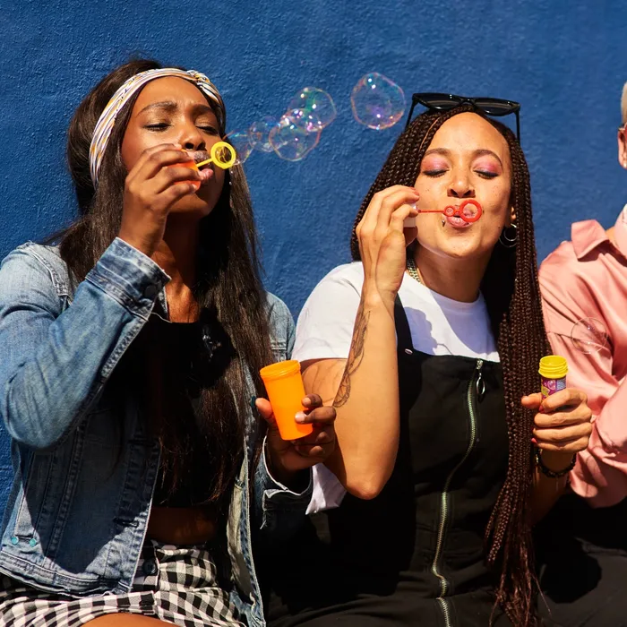 Cropped shot of three attractive young women sitting against a blue wall together and bonding by blowing bubbles