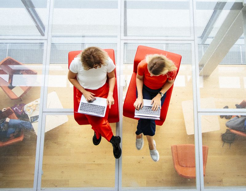 Top-down view of two women sitting with laptops in a contemporary office filled with natural light.Colorful Human