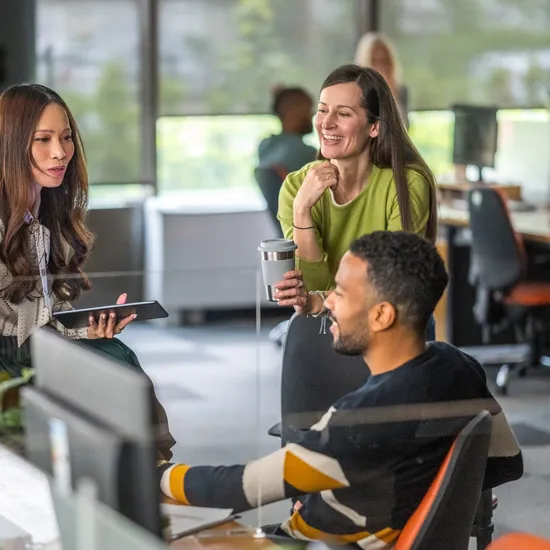 A group of diverse professionals collaborate enthusiastically in a modern office, exchanging ideas with digital devices on hand, while dressed in smart casual attire.
