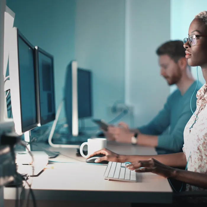 Closeup side view of mid 20's blond black woman doing her design project on a computer. She might be a software developer as well.  She's sitting in front of a desktop computer and sipping a coffee while working on a computer.