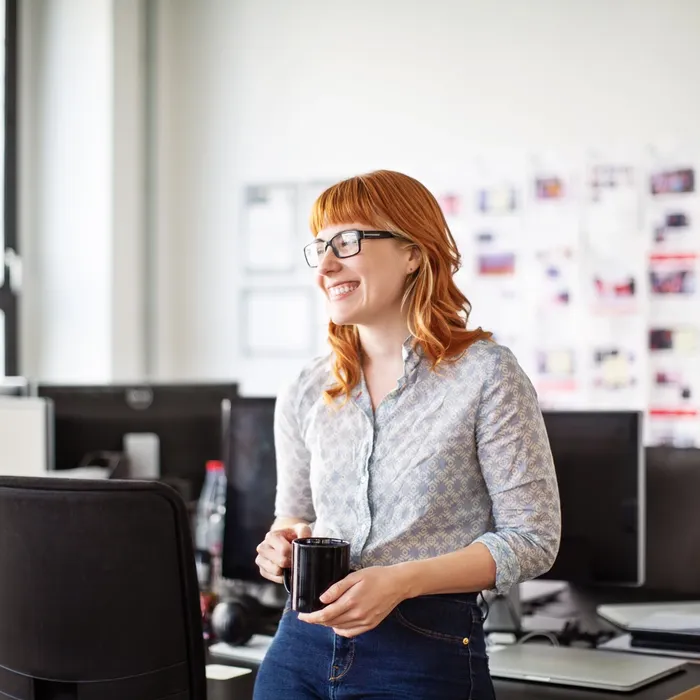 Smiling young businesswoman standing by her desk with cup of coffee. Female professional having a coffee break in office.