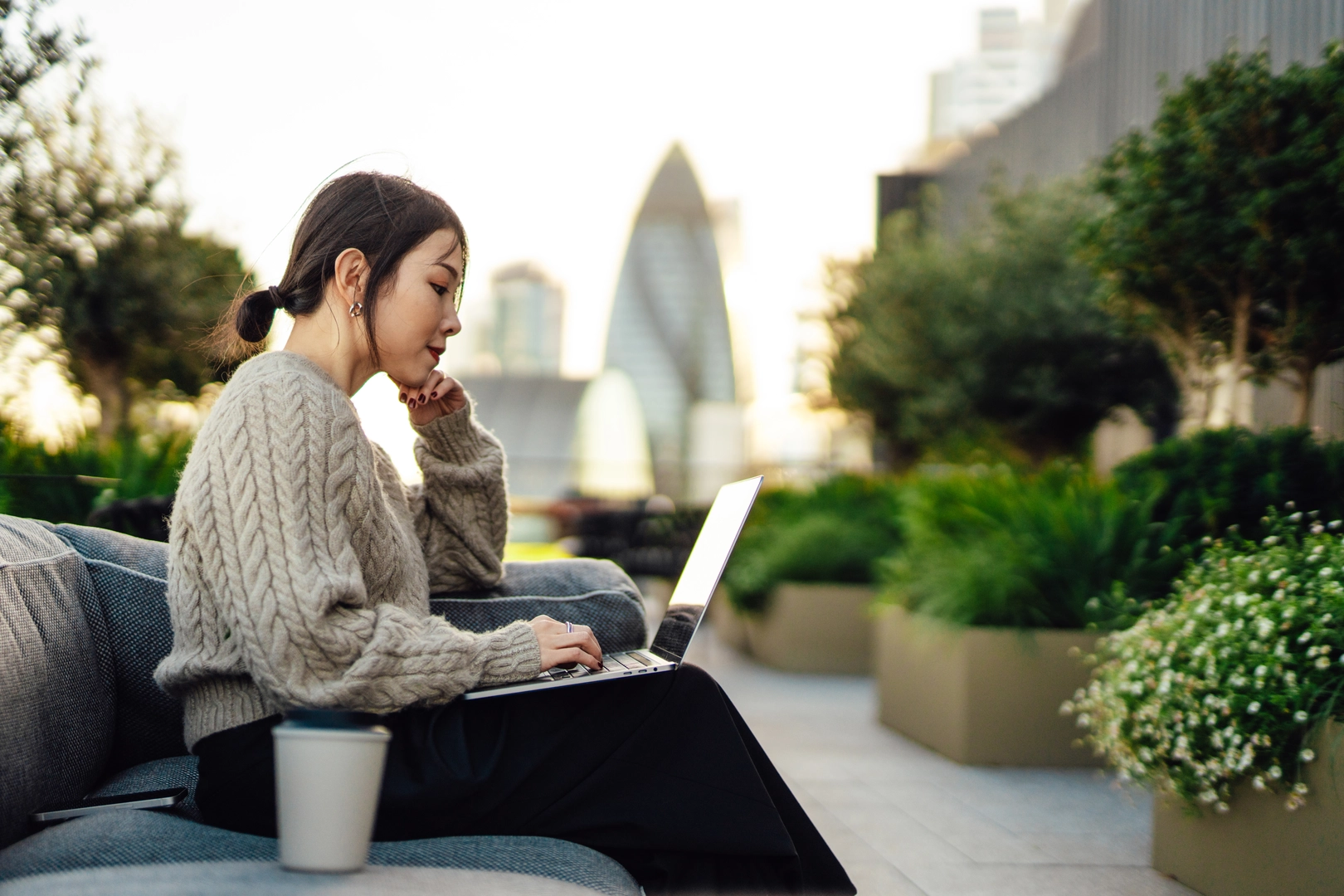 Young Asian businesswoman working on laptop outdoors, sitting on the bench in rooftop garden, surrounded by green plants. Looking at laptop. Working outdoors with technology. Remote working concept. Enjoying a cup of coffee in the middle of a work day. Online studying / e-learning concept.