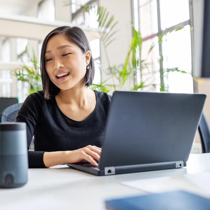 Asian businesswoman talking to virtual assistant at her desk. Female professional working on laptop and talking into a speaker.