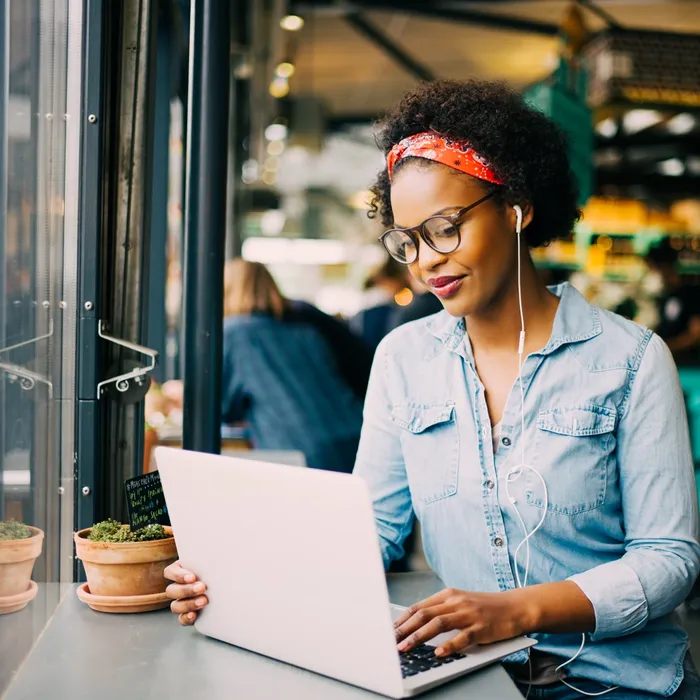 Focused young African woman sitting alone at a counter in a cafe working on a laptop and listening to music on earphones