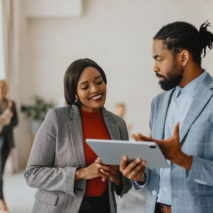 Business professionals collaborating and discussing work on a tablet in a modern office environment with colleagues in the background.Colorful Human