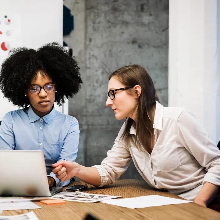 A black and a caucasian young woman are sitting in a business meeting in a modern office.They discuss something over papers and a laptop while one of them is pointing at the computer screen. Both are wearing glasses and pie charts are visible in the background.