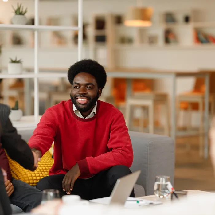 A diverse group of professionals engaged in a collaborative business meeting in a modern coworking space. A smiling Black man in a red sweater warmly shakes hands with a businesswoman in a dark blazer, symbolizing partnership and cooperation. Another White woman with short blonde hair observes the interaction with interest. The setting is a stylish, well-lit office space with contemporary decor, fostering a welcoming and productive environment.Colorful Human