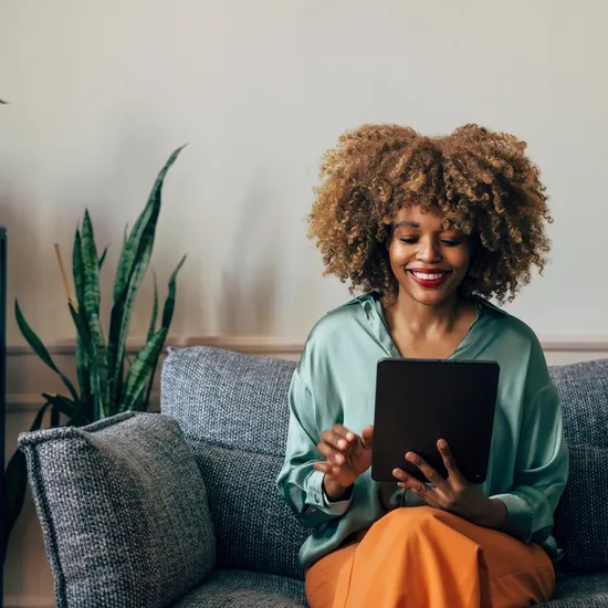 Cheerful young woman with curly hair enjoying digital content on a tablet while sitting comfortably on a couch.