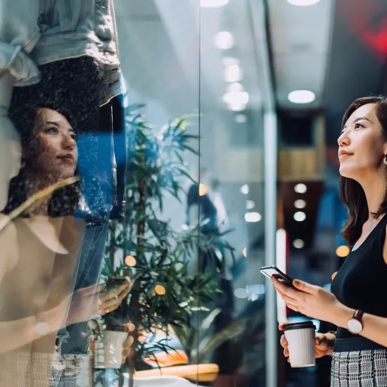 Cheerful young Asian woman holding a cup of coffee, checking her smartphone while standing outside a boutique looking at shop window in the evening in the city