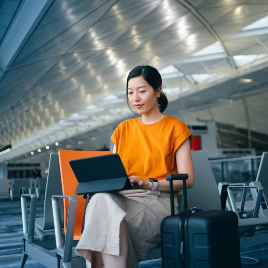 Young Asian businesswoman working with digital tablet while waiting for her flight in airport terminal, a suitcase by her side. Young Asian woman on business trip. Lifestyle and technology. Travel and vacation concept. Business on the go
