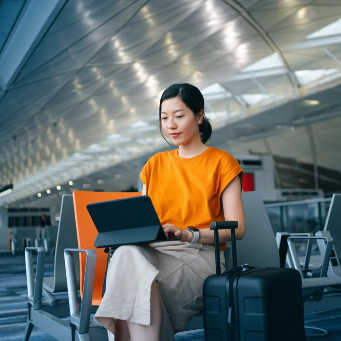 Young Asian businesswoman working with digital tablet while waiting for her flight in airport terminal, a suitcase by her side. Young Asian woman on business trip. Lifestyle and technology. Travel and vacation concept. Business on the go