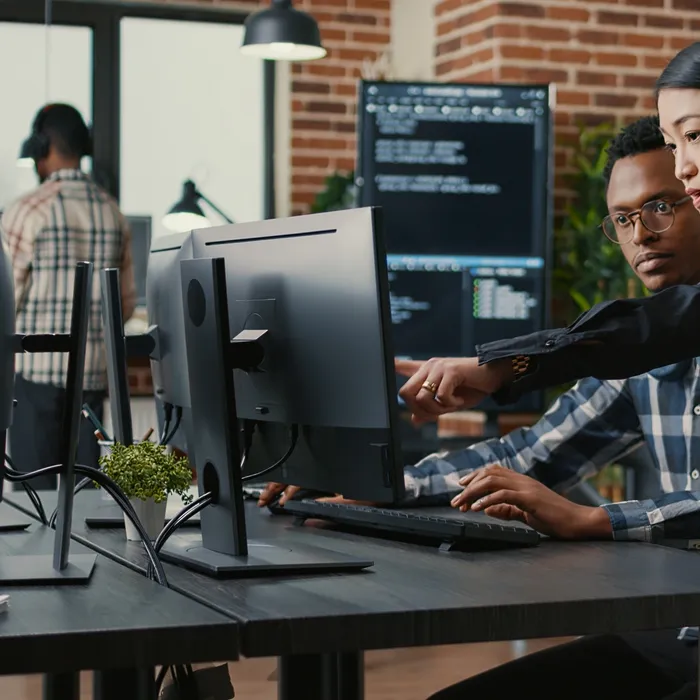 Two programers holding laptop with coding interface walking towards desk and sitting down talking about online cloud computing. Software developers team 
