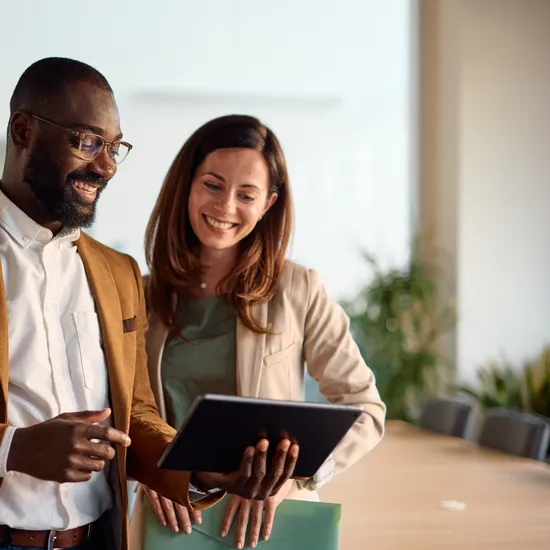 Two happy business colleagues engaging with a digital tablet and discussing various projects while standing in a bright, modern office environment, showcasing teamwork and collaborationColorful Human