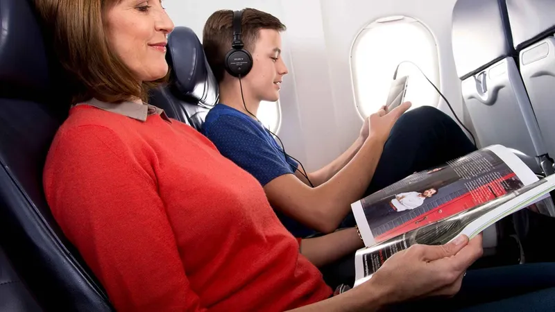 A woman in a red blouse is looking at a magazine and a teenager is looking at a tablet while they are on a plane