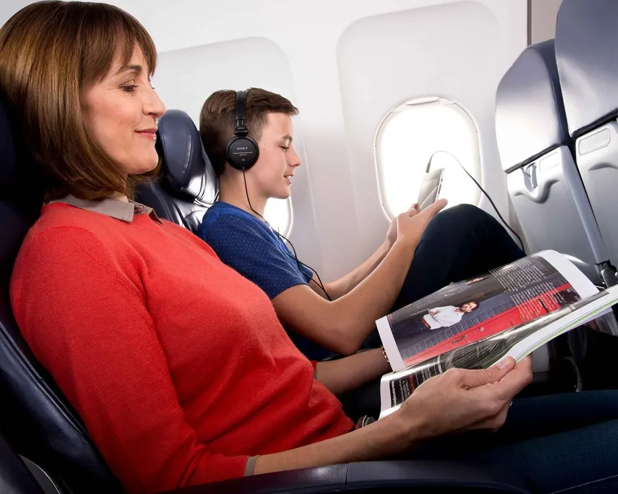 A woman in a red blouse is looking at a magazine and a teenager is looking at a tablet while they are on a plane