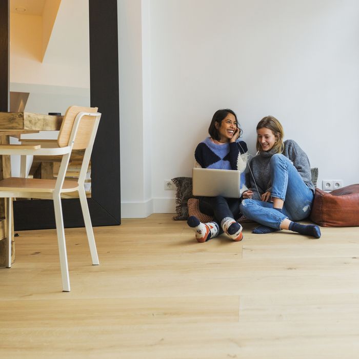 Two young women looking at a laptop having fun, leaning against a wall. Sitting in a living room on the ground.