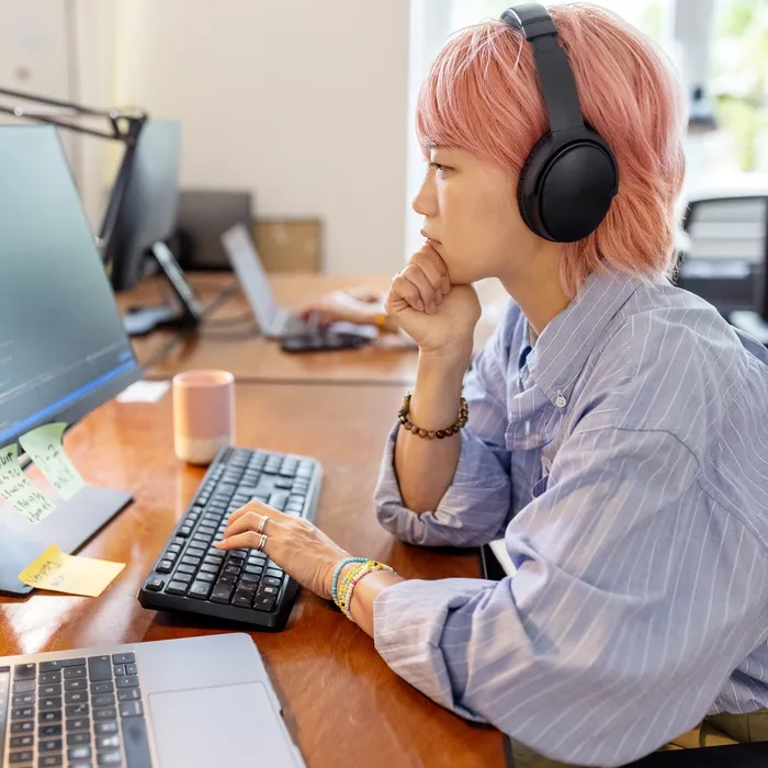 Side view of a woman computer programmer working at her desk at a startup company. Businesswoman wearing a headset looking at computer monitor and thinking while coding at coworking office space.